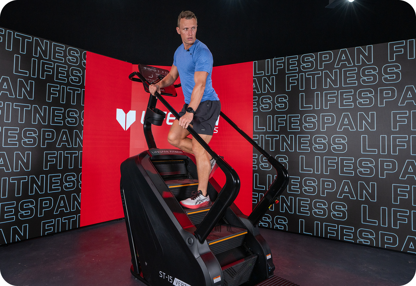 Man exercising on a stair climber in a gym, wearing a blue shirt and black shorts. Background has branding with Lifespan Fitness, creating a modern and energetic atmosphere.