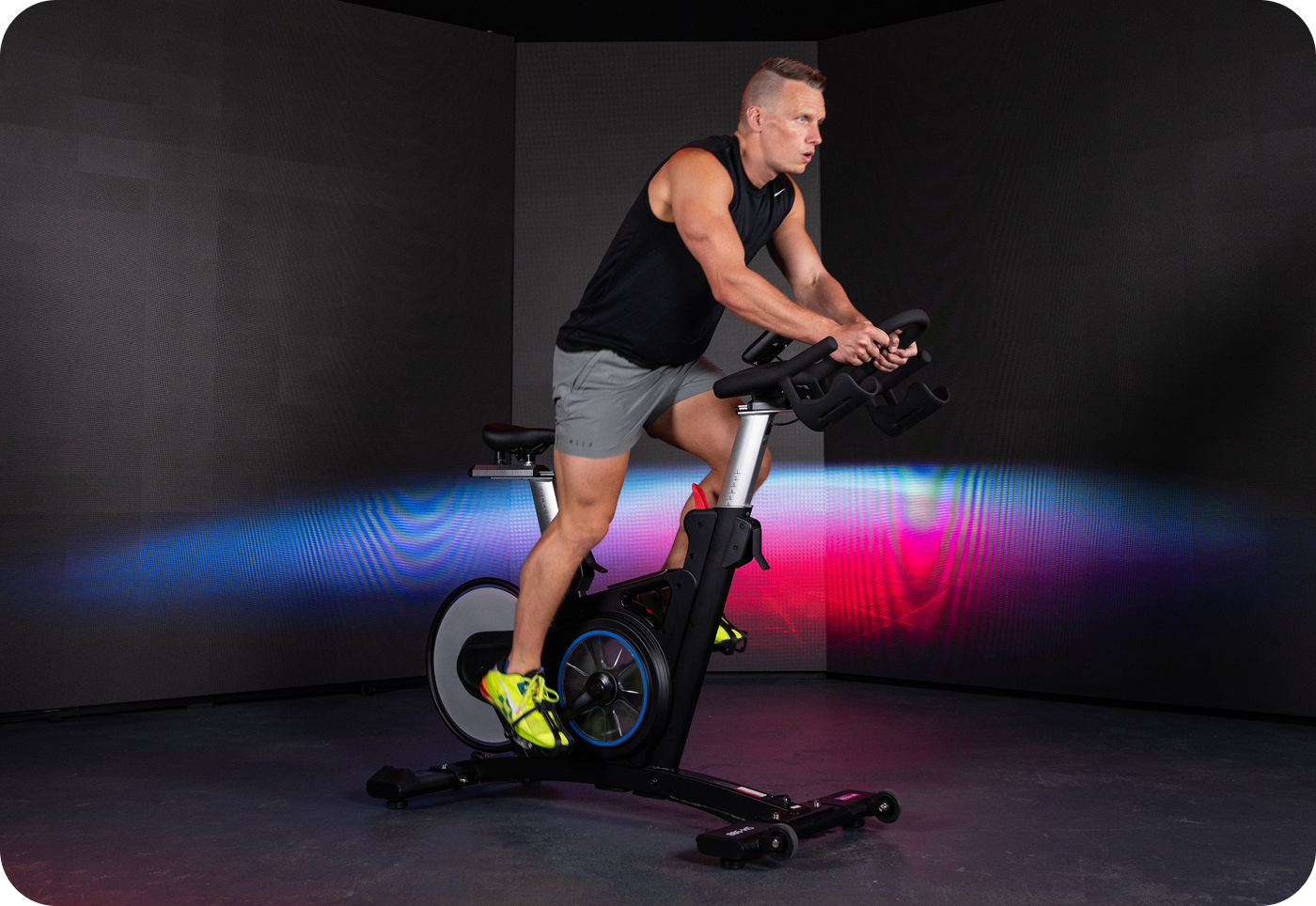 A man in workout attire uses an exercise bike in a home gym. He's focused on the machine. The room is well-lit with fitness equipment, creating a motivating atmosphere.