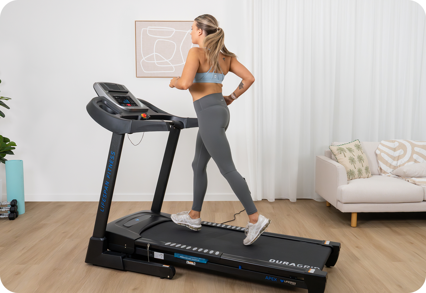 A woman jogs on a treadmill in a bright room with light wooden floors and a comfy sofa. She wears a sports bra and leggings, conveying an active, focused tone.