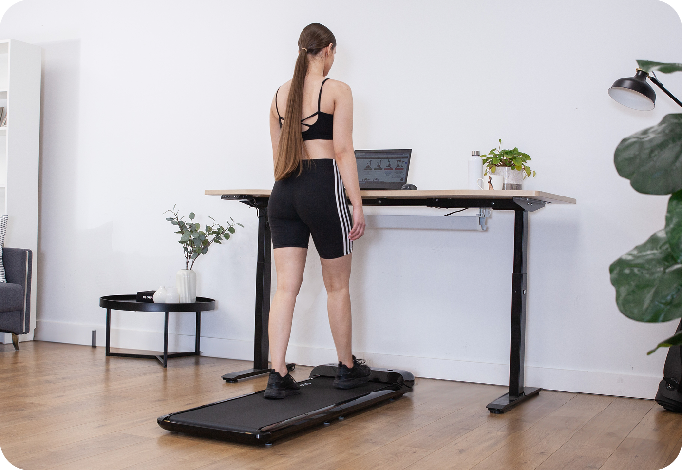 A woman in workout attire uses a treadmill desk at home. She's focused on a laptop. The room is bright with plants, creating a productive atmosphere.
